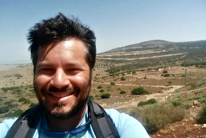 Matt Adler smiles at the camera while wearing backpack and taking a selfie in front of an Israeli desert setting