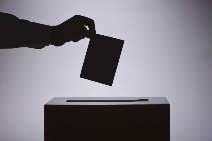 Black and white shadowy image of a hand dropping a ballot into a box