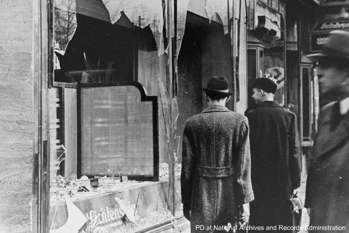 Black and white image of Germans looking at broken storefront windows the day after Kristallnacht