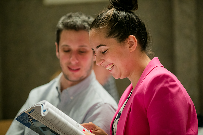 A person smiling while reading from a textbook, while another person follows along.