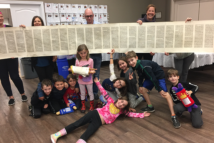 an image of a group of adults holding an opened Torah scroll while a group of students pose in front of the Torah