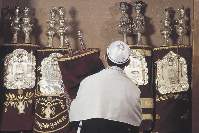 an image of a boy standing in front of four Torah scrolls while holding one and wearing a yamuka and tallit