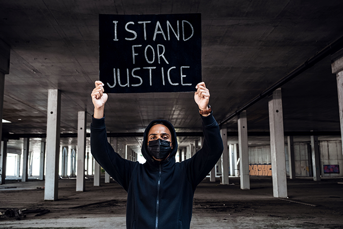 Photo of a masked man standing in anbandoned building holding a sign that say "I Stand for Justice."