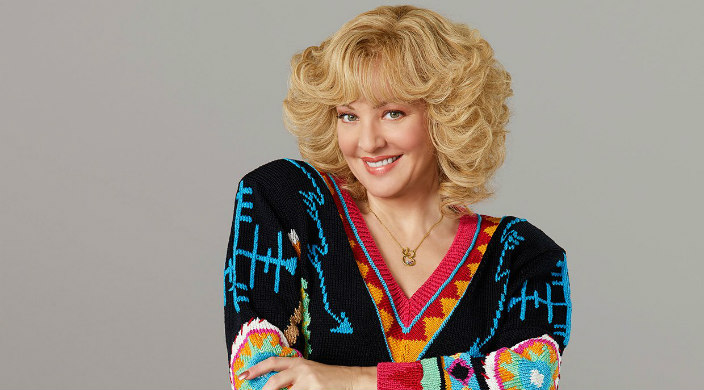 Blond woman from the 1980s with big hair and a colorful sweater with her arms crossed toward the camera