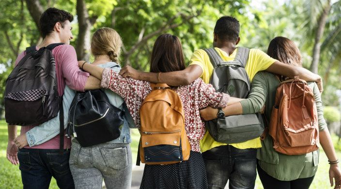 A group of young people with their backs to the camera and their arms around one another while wearing backpacks