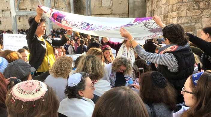 Women at the Kotel holding a prayer shawl over other women who are reading from the Torah 