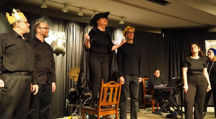 Individuals dressed in all black costumes acting out the Purim story on a small stage