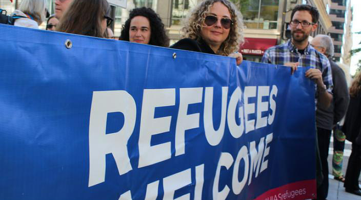 Two individuals holding a large banner sign that reads REFUGEES WELCOME