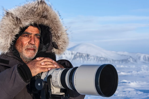 Amos Nachoum wearing a thick winter jacket and holding a large camera with an Arctic landscape visible behind him