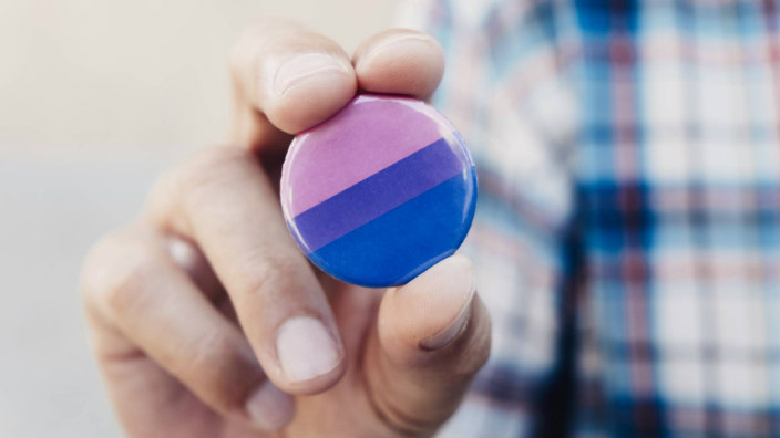 Close up of a persons fingers holding a button bearing the colors of the bisexuality pride flag 