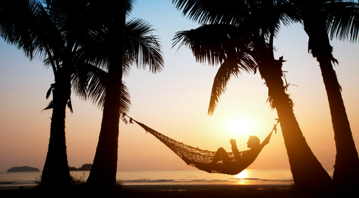 Silhouette of a person reading in a hammock strung between two palm trees on the beach at sunset