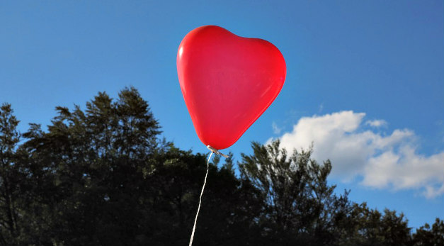 Red heart balloon rising into a blue sky over treetops