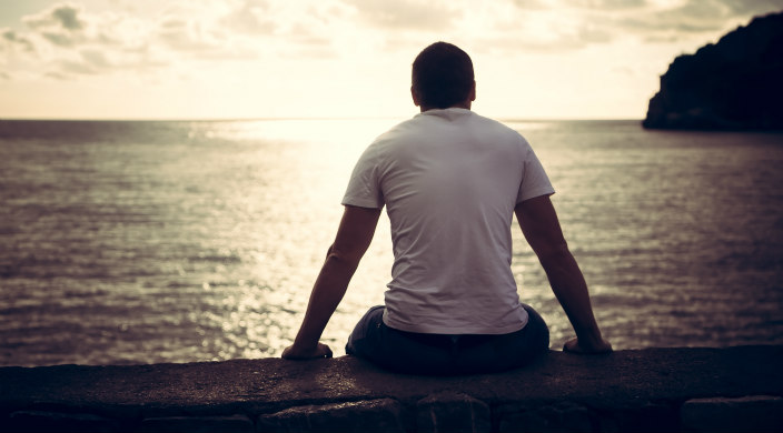 Figure from behind in tee-shirt and jeans, sitting on concrete wall and looking out at a body of water on which the sun is shining through clouds
