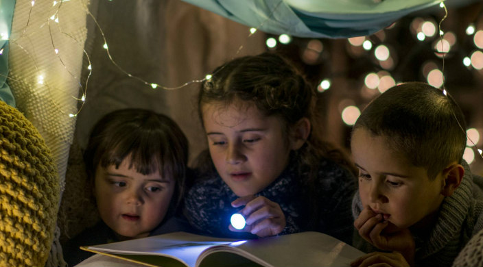 Three children using a flashlight to read a book inside a dark pillow fort 