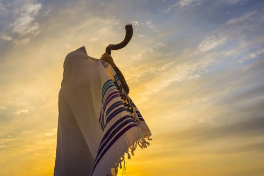 View from behind of a person blowing a shofar into the sunset while wearing a prayer shawl that covers their head and back
