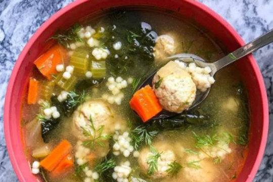 Red bowl full of Italian wedding soup with veggies meatballs and Israeli couscous