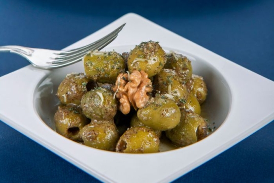 White square bowl containing green olives marinated in herbs with a walnut on top 