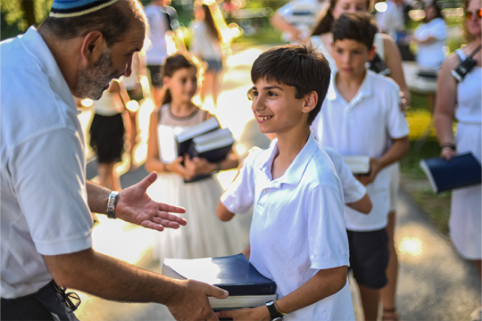An adult man waring a yarmulke speaks to a young boy holding books
