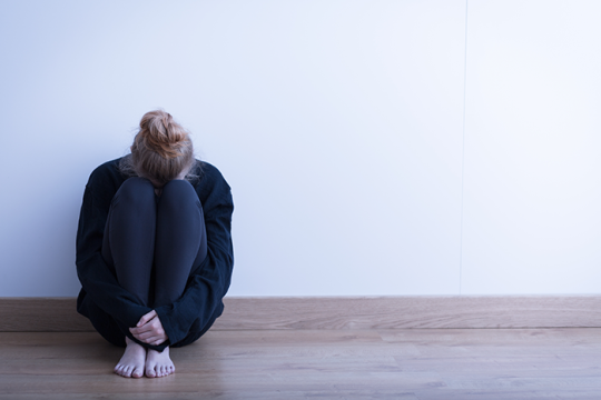 an image of a woman sitting by herself against a white wall with her legs up and her head resting on her knees