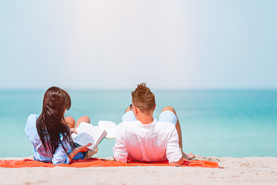 Photo of a man and woman laying on a blanket on the beach, the woman is holding a book