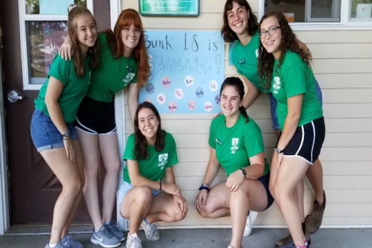 Smiling camp staffers pose in front of a bunk sign