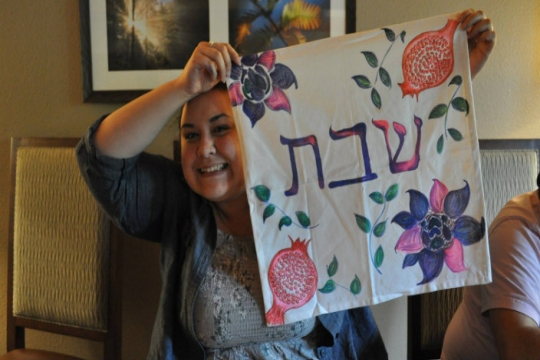 Amanda Ryan smiles while holding up a challah cover with Hebrew words and a hand drawn pomegranate