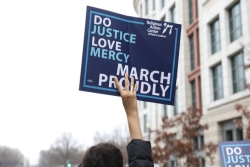 Hand holding a sign in the air that reads DO JUSTICE LOVE MERCY WALK PROUDLY