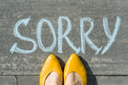 Womans feet and pointed yellow dress shoes standing in front of the word SORRY written on the pavement in chalk