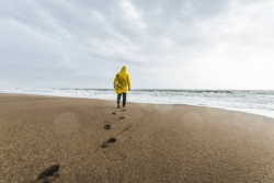 A person in a yellow jacket faces the ocean while standing at the edge of a beach with a line of footprints in the sand