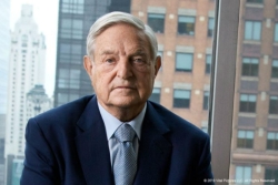 Headshot of George Soros wearing a blazer and blue tie in front of a window with high rise buildings in background
