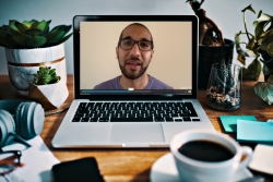 Laptop on a desk surrounded by items like coffee and headphones and showing a young man speaking on screen