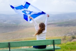 Young girl siting on a bench overlooking a scenic view of Israel while holding an Israeli flag above her head