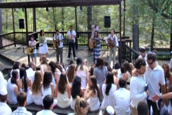 Songleaders dressed in white playing guitar and singing on an outdoor stage at URJ Camp Newman looking out over Shabbat services attended by campers