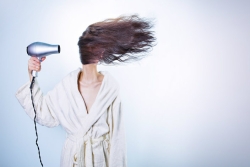 Woman in a white robe with her hair blowing straight across her face as she aims a hairdryer at it