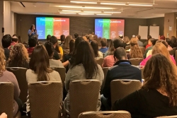 Shot from the back of a large room where a woman is speaking at the front and giving a presentation