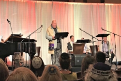 Cantor Steven Weiss stands on a stage to give a dvar Torah before a sea of Jewish musicians wearing kippot and tallitot at a worship service 
