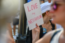 Sign reading "Love Thy Neighbor" held up against a blurred green background, at a protest