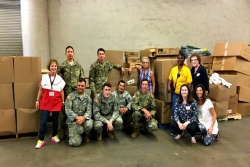 Group of smiling volunteers including some in military uniforms posing with large boxes in a cement warehouse 
