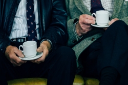Close ups of two older men sitting next to one another while holding cups of coffee 