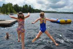 View from behind of two young girls in bathing suits jumping into a lake with a thick forest along the banks