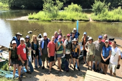 Group photo of about two dozen adults standing on a river bank wearing kayaking gear on a sunny day