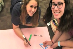 Two female students sitting on the ground and smiling up at at the camera while drawing Stars of David on posterboard
