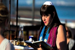 Female rabbi in her twenties or thirties dressed in a prayer shawl and kippah while reading from a prayerbook outdoors