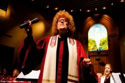 Closeup of a female rabbi with curly blond hair leading a congregation from the bimah with her arms open wide while wearing a prayer shawl 