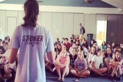 Teen girl facing away from the camera in a shirt that reads STRONGER in front of a seated audience of other teens