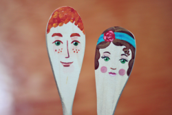 two spoon puppets, one depicting a man and the other a woman. The man has red hair and a smile. The woman has brown bob haircut, a blue headband, blush, and pink lipstick