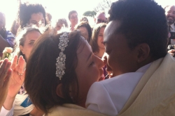 An interracial lesbian couple shares their first kiss on their wedding day with their friends and family cheering in the background