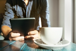 Man sitting at a wooden table with a cup of coffee next to him as he reads from an iPad