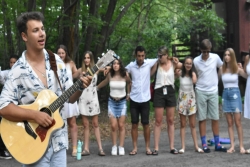 Songleader strumming a guitar in the foreground with people gathered in a circle around him in the background