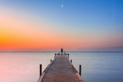 man on end of dock with sunset and moon